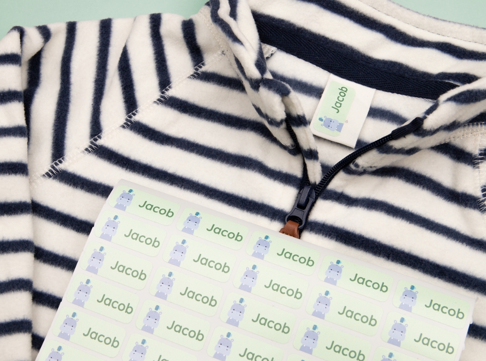 Étiquettes nominatives : dois-je choisir des étiquettes autocollantes ou thermocollantes? 2 Planche d’étiquettes autocollantes vertes avec motif d’hippopotame, posée sur un t-shirt rayé pour enfant