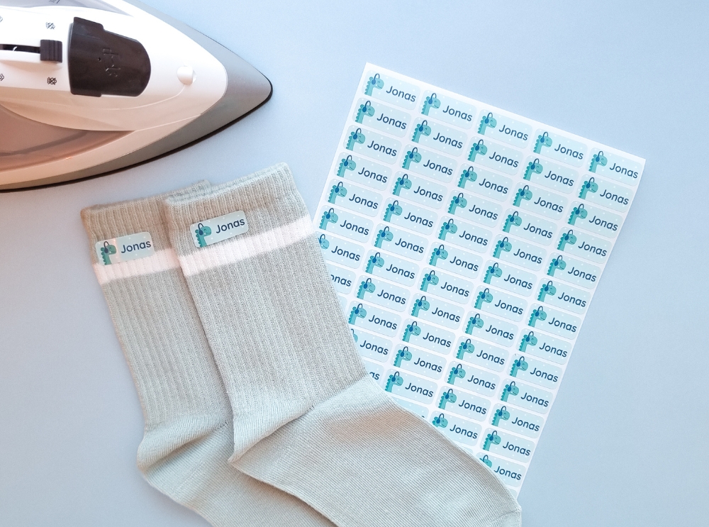 Étiquettes nominatives : dois-je choisir des étiquettes autocollantes ou thermocollantes? 3 Planche d’étiquettes thermocollantes avec dinosaure portant un casque audio, à côté de chaussettes étiquetées et d’un fer à repasser