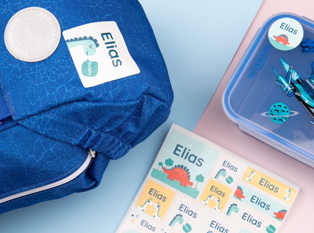 Étiquettes nominatives : dois-je choisir des étiquettes autocollantes ou thermocollantes? 4 Autocollants nominatifs colorés avec dinosaures sur un sac à dos, une boîte à lunch et une planche d’étiquettes